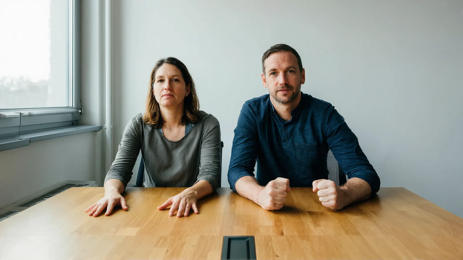 Two colleagues sit side by side at a meeting-room table with identically calm, neutral faces. The woman's hands rest flat and open on the table with fingers relaxed; the man's hands are closed into two tight fists.